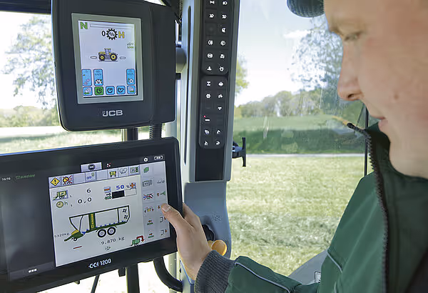Operatør betjener JCB-display og CCI 1200-monitor inne i KRONE høyladervogn med utsikt til mark.