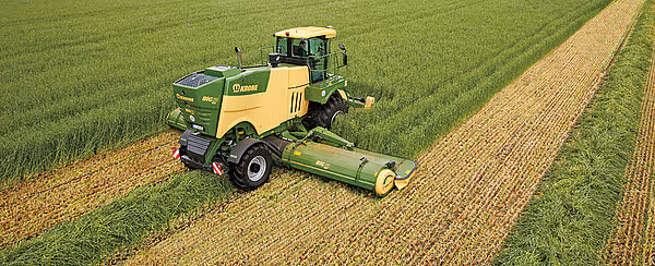 Luftfoto av Krone Big M 450 slåtterkondisjonerer som kutter gress i lange, rette striper på en grønn eng. Maskinen etterlater tydelige kjørespor og kuttet gress bak seg.