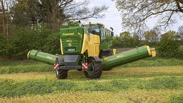 En KRONE Big M 450 slåmaskin arbeider på en eng med store trær i bakgrunnen. Maskinen har brede slåaggregater på hver side og slår høyt gras.