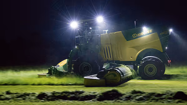 Krone slåmaskin i arbeid om natten med kraftig arbeidsbelysning. Maskinen kaster opp støv og gress under slåttearbeidet.
