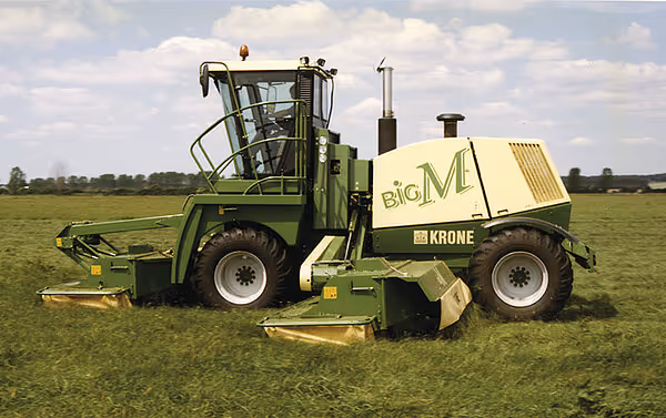 En eldre modell KRONE Big M slåmaskin på et åpent jorde under skyet himmel. Maskinen har et kompakt design og er utstyrt med frontmontert slåaggregat.