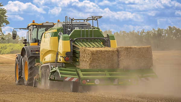 Krone Big Pack ballepresse i aksjon på åker med støv som virvler opp bak maskinen. Den grønn-gule ballpressen produserer store firkantballer.