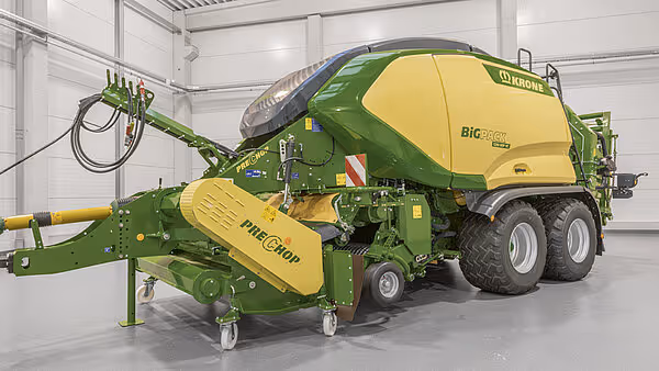 KRONE BigPack ballepresse med InteCrop pickup parkert innendørs, med grønn og gul fargeskjema. Maskinen har store hjul og er koblet til en grønn pickup-enhet.