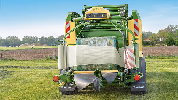 Bakside av KRONE Comprima Plus rundballpresse som står på eng med ferdig innpakket hvit rundball. Maskinen har karakteristisk grønn og gul design.
