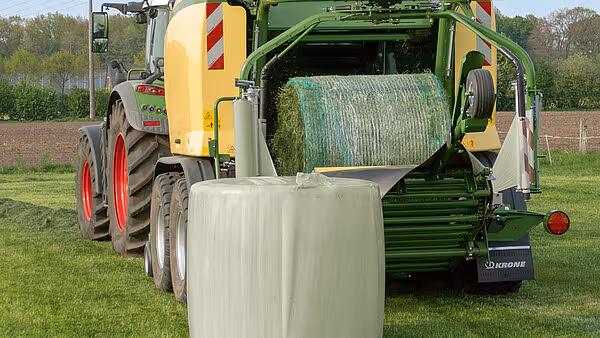 KRONE Comprima Plus rundballpresse i arbeid med traktor på bakgrunn. Maskinen pakker rundballer i hvit plastfilm på grønt jorde.