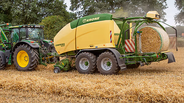 En traktor med KRONE Comprima Plus rundballpresse i arbeid på en stubbeåker, med en ferdig rundball synlig i pressa.