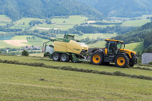 KRONE Comprima rundballpresse trukket av gul traktor på slåttemark i kupert landskap med skog og fjell i bakgrunnen.