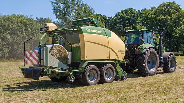 KRONE Comprima rundballpresse trukket av grønn traktor på stubbeåker, med ferdig rund halmballe synlig bak maskinen.