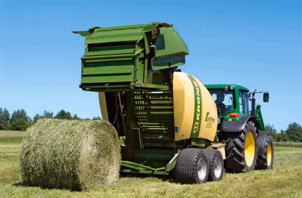KRONE Fortima rundballpresse med åpen bakkant som slipper ut en ferdig rundball. Pressen er koplet til en grønn traktor på en høyåker.