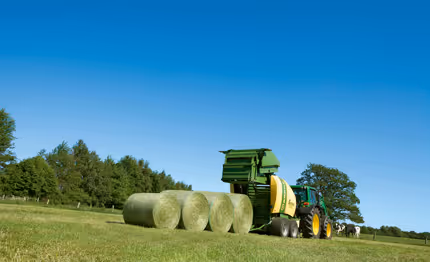 Krone Fortima rundballpresse i arbeid på eng med ferdigpressede rundball i bakgrunnen. Blå himmel og grønt landskap rundt.
