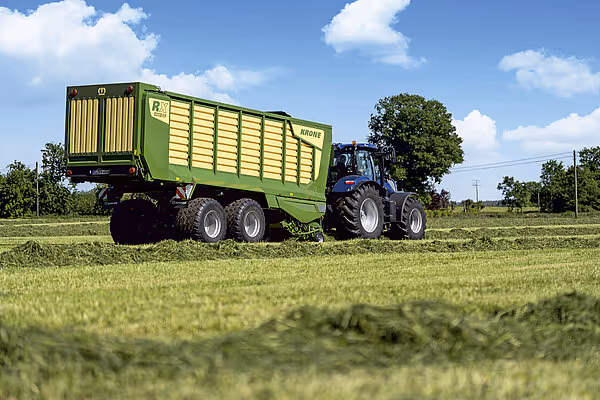 En blå traktor trekker en grønn og gul Krone RX fôrvogn lastet med gras på en eng.