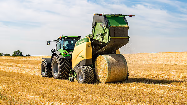 KRONE Varipack Plus rundballpresse trekkes av en grønn traktor på en høstet kornåker. Maskinen har akkurat produsert en stor rundball.