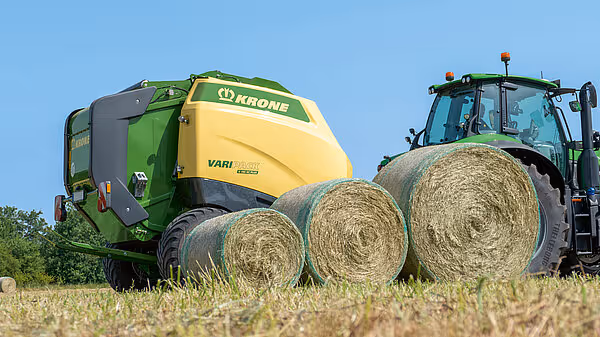 KRONE Varipack Plus rundballpresse med traktor står ved siden av tre ferdiglagde rundaller på en stubbeåker. Maskinen er gul og grønn i farger.