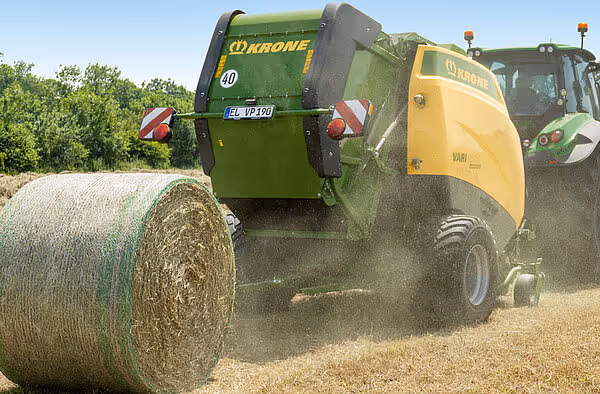 KRONE Varipack Pro rundballpresse i arbeid på kornstubbing, med ferdig rundball som trilles ut av maskinen. Støv virvler opp under arbeidet.