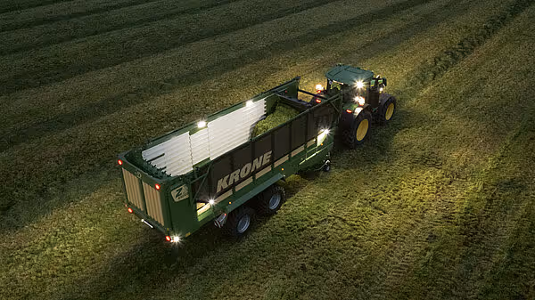 Grønn KRONE lastekar trukket av traktor under nattlig høsting på eng, med arbeidsbelysning.