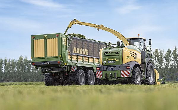 KRONE selvgående forhakser med gul kabin og grønn oppsamlingsvogn arbeider på en grønn eng under blå himmel.
