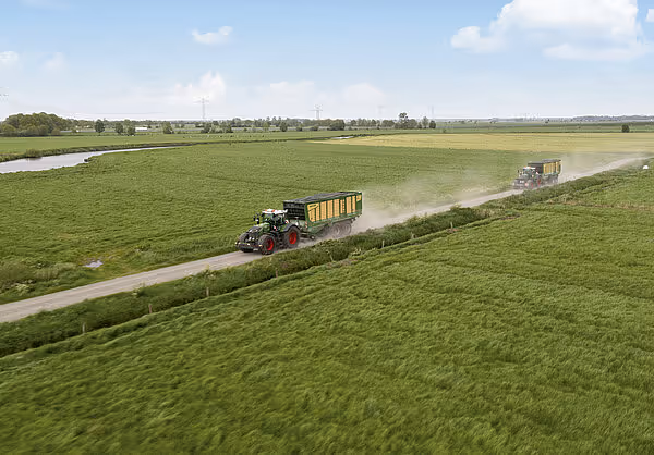 Luftfoto av traktor med KRONE oppsamlingsvogn som transporterer høstet gras langs en grusveien i grønt landskap.
